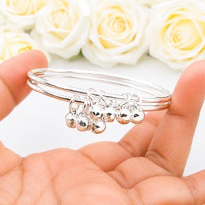 May include: A silver-coloured bangle bracelet featuring two thin, smooth bands. Attached to the bands are several small, round, reflective silver-coloured bell charms that hang from individual jump rings, creating a clustered, metallic appearance.