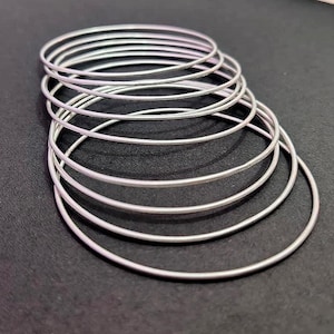Op de afbeelding: Een stapel zilverkleurige armbanden. De dunne, ronde armbanden zijn in een licht gebogen formatie gerangschikt, met verschillende zichtbaar. De armbanden zijn gemaakt van een glanzend, metalen materiaal dat licht reflecteert. De achtergrond is donker en effen.