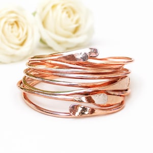 Op de afbeelding: Een stapel van meerdere koperkleurige metalen armbanden met een gehamerde, reflecterende afwerking. Elke individuele armband heeft een glad, afgerond uiteinde en de set is gerangschikt in een cirkelvormige, overlappende formatie.