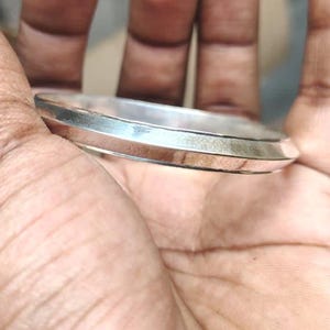 May include: A silver bangle bracelet with a flat top and angled edges. The bracelet is circular and seems to be made of metal. The jewellery is shown on a person's hand.