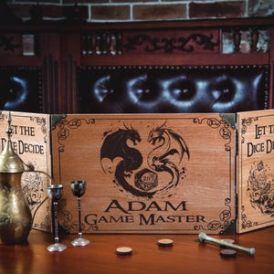 May include: A wooden three-panel game master screen featuring laser-engraved dragon illustrations, a twenty-sided die graphic, and the text "Let the Dice Decide" and "Adam Game Master." A brass-colored metal pitcher and two small metal goblets are present.