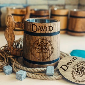 Puede incluir: Jarra de cerveza de madera con un asa tallada y el nombre "DAVID" grabado en la parte delantera. La jarra tiene un interior de metal y un diseño decorativo. También hay un posavasos a juego y cubitos de hielo.