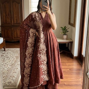 May include: A terracotta-coloured, sleeveless dress with a V-neck and a flowing skirt. The outfit includes matching trousers and a shawl with detailed gold embroidery. The ensemble is paired with white flats. The style is elegant and traditional.
