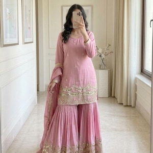 May include: A light pink Sharara suit with gold embroidery. The long-sleeved top and wide-legged pants feature intricate gold detailing along the hem and cuffs. The outfit includes a matching dupatta and is paired with white shoes.