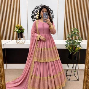 May include: A pink, tiered formal dress with gold embroidery. The dress has long sleeves with gold detailing and a matching scarf. The dress is reflected in a gold-framed mirror.