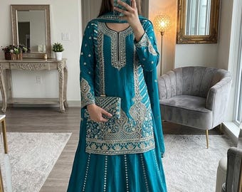 Puede incluir: Un conjunto de túnica de manga larga y falda hasta el suelo en color verde azulado, con intrincados bordados plateados y detalles de espejos. El conjunto incluye un dupatta a juego y un pequeño bolso de mano rectangular.