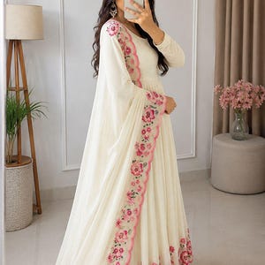 May include: A floor-length cream-colored dress with long sleeves and a matching shawl. The garment features a scalloped edge adorned with pink floral embroidery and green leaf accents along the hem and the border of the draped shawl.