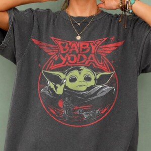 May include: Dark gray t-shirt featuring a graphic of Baby Yoda in a red circle with the text "BABY YODA". The shirt has short sleeves and a relaxed fit. The bottom left corner of the shirt has the text "PEPPER COMFORT COLORS".