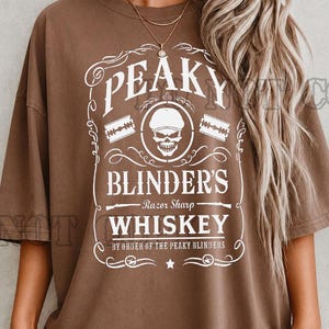 May include: Brown t-shirt with a white graphic design. The design features the text "PEAKY BLINDER'S WHISKEY" with a skull and razor blades. The shirt has a vintage, worn look.