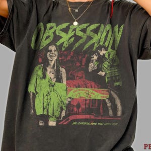 Könnte beinhalten: Ein anthrazitfarbenes Kurzarm-T-Shirt mit einem Grafikdruck, der den Text "OBSESSION" in Grün zeigt. Das Design umfasst eine Person in einem grünen Hemd und ein Paar, mit dem Text "BE CAREFUL WHO YOU WISH FOR" am unteren Rand.