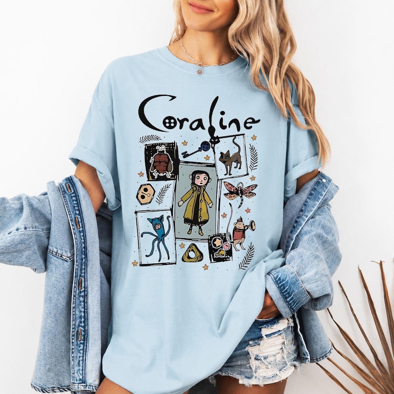 May include: A light blue t-shirt featuring a graphic print with the text Coraline. The design includes illustrations of the character Coraline, a black cat, a key, a dragonfly, and other icons arranged in a collage style with various small stars.