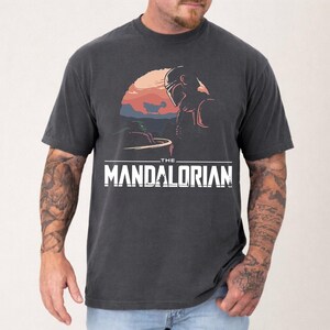 Pu&ograve; includere: T-shirt grigio scuro con una grafica di The Mandalorian. Il design include una scena del tramonto con il Mandaloriano e Grogu, e il testo "THE MANDALORIAN" in lettere bianche.