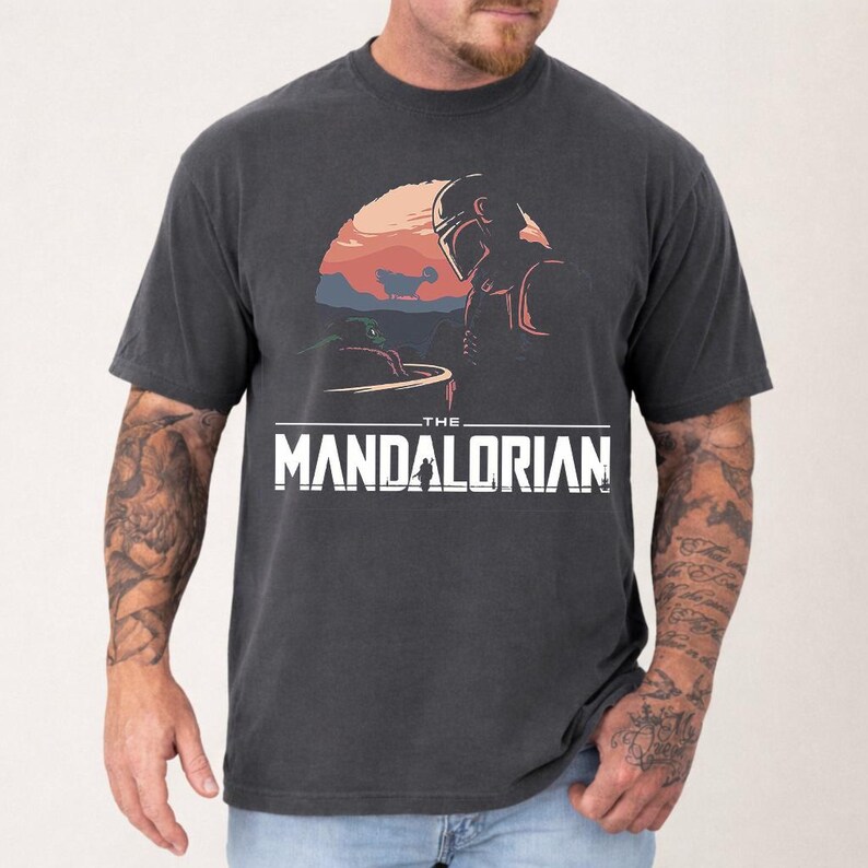 Pu&ograve; includere: T-shirt grigio scuro con una grafica di The Mandalorian. Il design include una scena del tramonto con il Mandaloriano e Grogu, e il testo "THE MANDALORIAN" in lettere bianche.