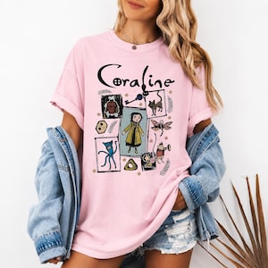 May include: A light pink short-sleeved t-shirt featuring a graphic print with the text Coraline. The design includes illustrations of the character Coraline, a black cat, a key, a dragonfly, and other small icons arranged in a rectangular layout.