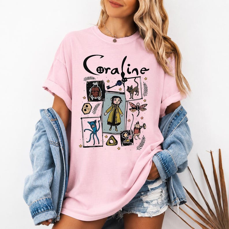 May include: A light pink short-sleeved t-shirt featuring a graphic print with the text Coraline. The design includes illustrations of the character Coraline, a black cat, a key, a dragonfly, and other small icons arranged in a rectangular layout.