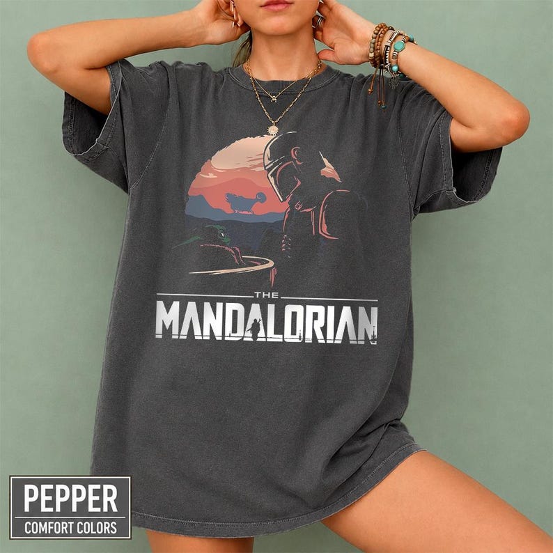 Pu&ograve; includere: T-shirt grigio scuro con una grafica di The Mandalorian con uno sfondo di tramonto. Il testo "THE MANDALORIAN" &egrave; in bianco. La maglietta ha anche il testo "PEPPER COMFORT COLORS" in una scatola bianca.