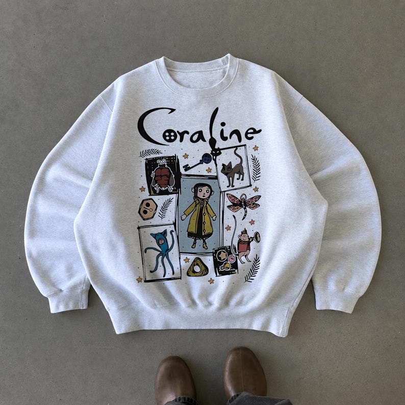 May include: A light grey crewneck sweatshirt featuring a graphic print with the text Coraline. The design includes a central illustration of a character in a yellow coat surrounded by smaller drawings of a key, a cat, a dragonfly, and other icons.