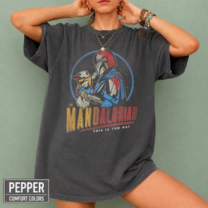 Pu&ograve; includere: T-shirt grigio scuro con grafica circolare di The Mandalorian e Grogu. Il design include il testo "THE MANDALORIAN" in giallo e "THIS IS THE WAY" in rosso. La maglietta &egrave; in stile comfort colors.