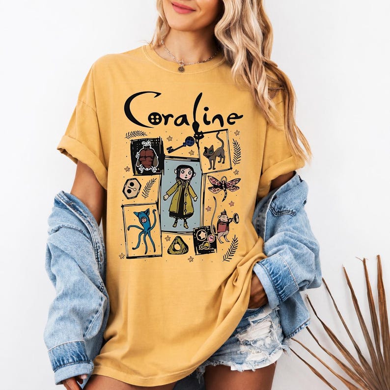 May include: A mustard yellow short-sleeve t-shirt featuring a black graphic print with the text Coraline. The design includes a central illustration of a character in a yellow coat surrounded by smaller drawings of a cat, a key, and creatures.