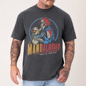Pu&ograve; includere: T-shirt grigio scuro con una grafica di The Mandalorian e Grogu, all'interno di un cerchio blu. Il design include il testo "The Mandalorian" e "This is the Way" in stile vintage.