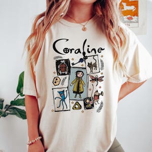 May include: A cream-coloured short-sleeve t-shirt featuring a graphic print with the text Coraline. The design includes a central illustration of a character in a yellow raincoat surrounded by various icons like a key, a cat, and a dragonfly.