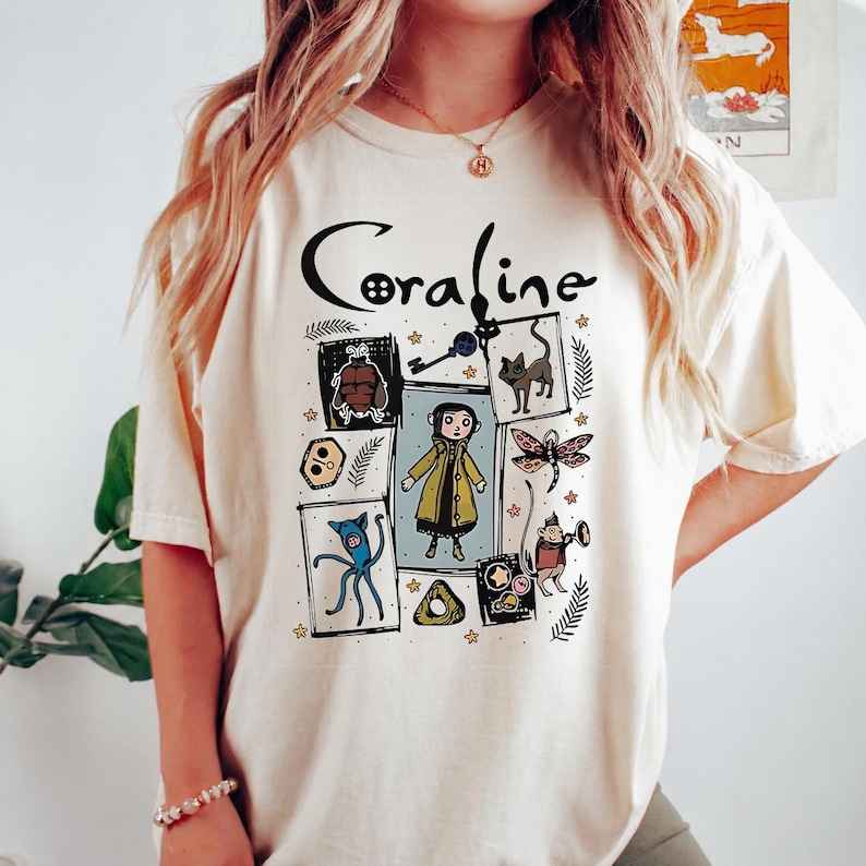 May include: A cream-coloured short-sleeve t-shirt featuring a graphic print with the text Coraline. The design includes a central illustration of a character in a yellow raincoat surrounded by various icons like a key, a cat, and a dragonfly.
