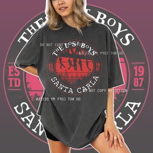 May include: Dark gray t-shirt with a circular graphic featuring the text "THE LOST BOYS SANTA CARLA 1987." The graphic includes silhouettes of figures and trees in red. The shirt has short sleeves and a relaxed fit.