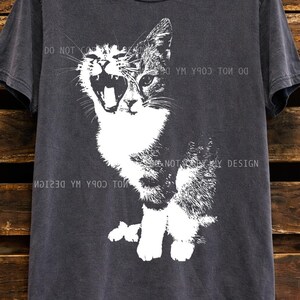May include: Dark gray t-shirt featuring a black and white graphic of a cat with its mouth open. The text "DO NOT COPY MY DESIGN" is printed on the shirt. The word "PEPPER" is printed at the bottom.
