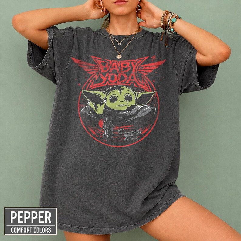 May include: Dark gray t-shirt featuring a graphic of Baby Yoda in a red circle with the text "BABY YODA". The shirt has short sleeves and a relaxed fit. The bottom left corner of the shirt has the text "PEPPER COMFORT COLORS".