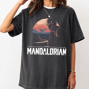 Pu&ograve; includere: T-shirt grigio scuro con una grafica di The Mandalorian e Grogu su uno sfondo di tramonto. Il testo "THE MANDALORIAN" &egrave; stampato in lettere bianche. La maglietta ha maniche corte e girocollo.