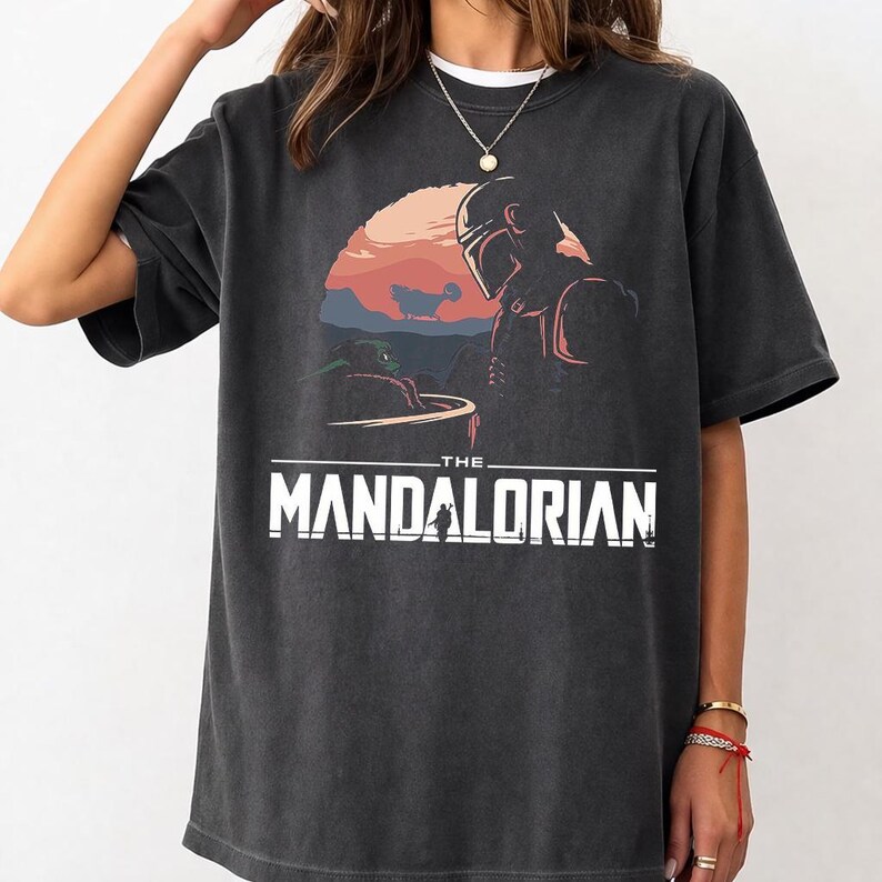 Pu&ograve; includere: T-shirt grigio scuro con una grafica di The Mandalorian e Grogu su uno sfondo di tramonto. Il testo "THE MANDALORIAN" &egrave; stampato in lettere bianche. La maglietta ha maniche corte e girocollo.
