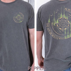 Puede incluir: Camiseta gris carbón con un gráfico circular en la parte delantera y trasera. El gráfico frontal es un diseño de brújula. El gráfico trasero presenta un diseño de brújula con el texto "ALWAYS TAKE THE SCENIC ROUTE".