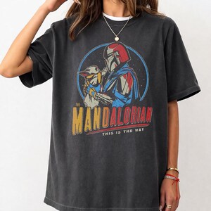 Vintage style oversized black t shirt featuring The Mandalorian holding Grogu with retro sci fi graphic design and This Is The Way slogan, unisex Star Wars inspired apparel.
