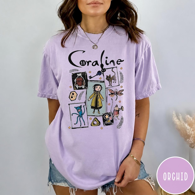 May include: A light purple short-sleeve t-shirt featuring the text Coraline at the top. The graphic includes a central illustration of a character in a yellow raincoat surrounded by smaller framed images of a cat, a key, a dragonfly, and other icons.