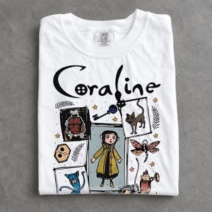 May include: A white short-sleeved t-shirt featuring the text Coraline in black lettering. The graphic includes a central illustration of a character in a yellow raincoat surrounded by various icons including a key, a cat, a dragonfly, and other figures.