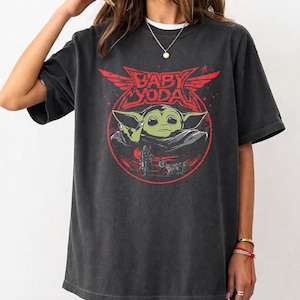 May include: Dark gray t-shirt featuring a red graphic of Baby Yoda with the text "BABY YODA" in a stylized font. The design includes a circular frame with a figure and vehicle. The shirt has a crew neck and short sleeves.