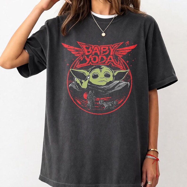 May include: Dark gray t-shirt featuring a red graphic of Baby Yoda with the text "BABY YODA" in a stylized font. The design includes a circular frame with a figure and vehicle. The shirt has a crew neck and short sleeves.