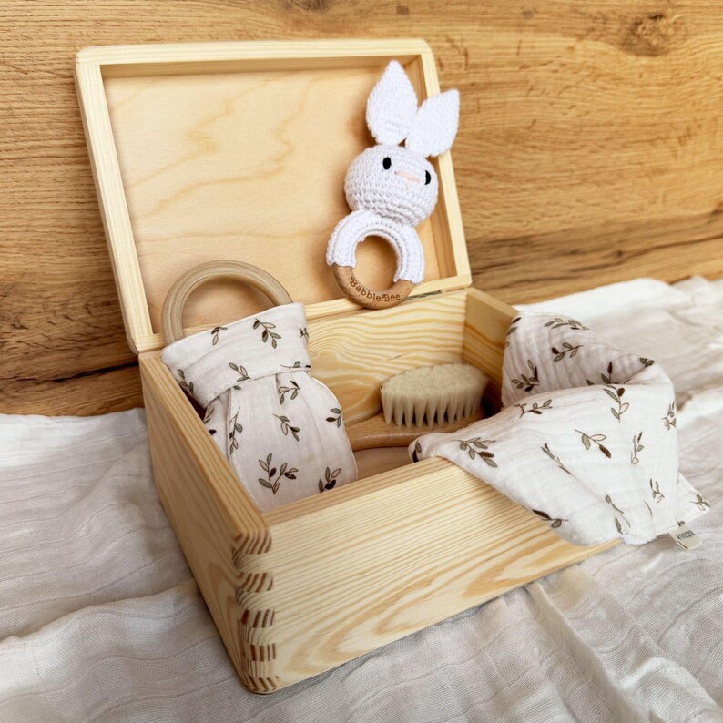 An open light wooden box containing a white crocheted bunny rattle, a wooden brush, and two folded white cloths with a green leaf pattern.