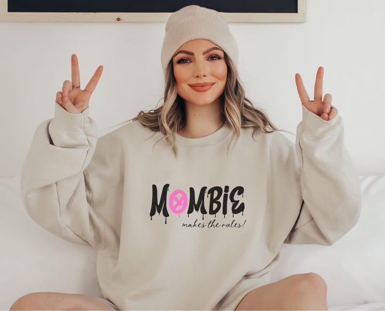 Könnte beinhalten: Beiges Sweatshirt mit dem Text "MOMBIE makes the rules!" in Schwarz mit rosa Akzenten. Das Sweatshirt wird mit einer beigen Mütze kombiniert. Das Model zeigt das Friedenszeichen.