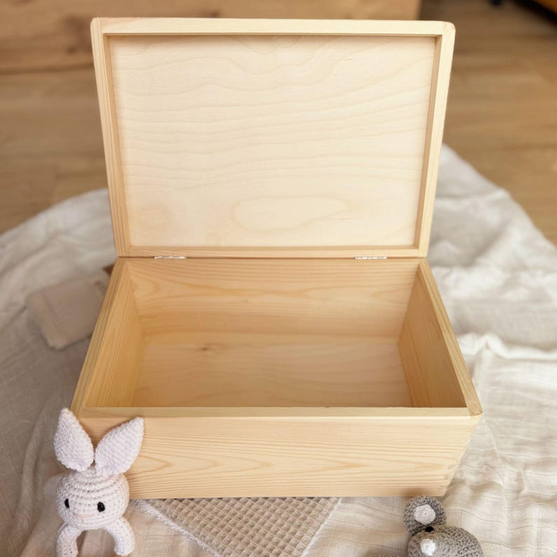 An open, light-colored wooden box with a hinged lid, showing its empty interior, placed on a white blanket next to a small white crocheted bunny toy.