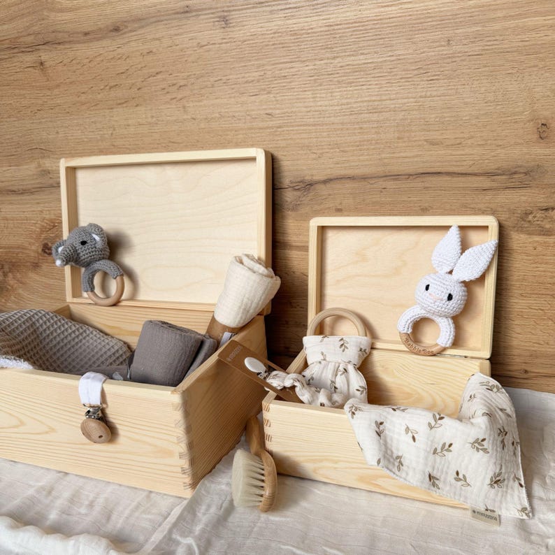 Two open light wooden boxes, one with a gray crocheted elephant rattle and gray cloths, the other with a white crocheted bunny rattle and white cloths with a green leaf pattern.