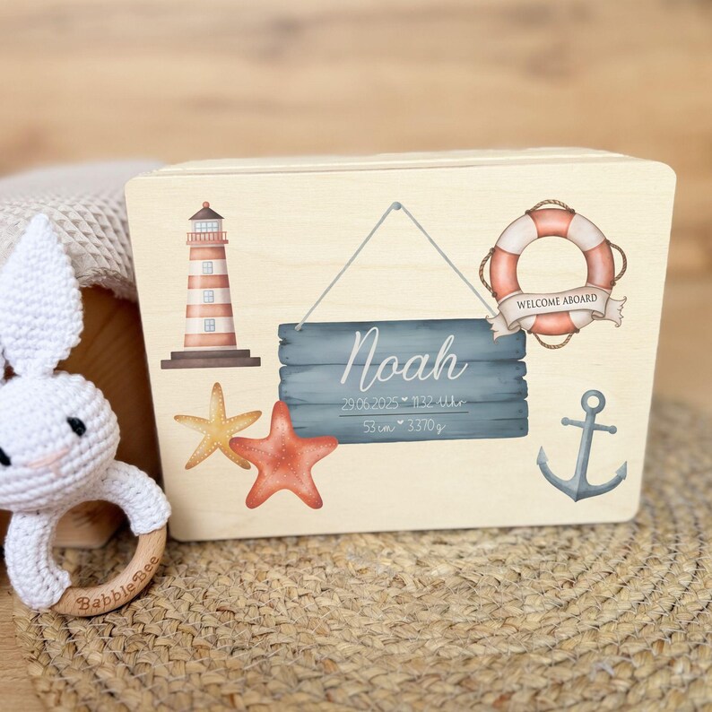 A closed light wooden box with a nautical design on its lid, featuring a lighthouse, starfish, a lifebuoy with "WELCOME ABOARD" text, an anchor, and a hanging sign with "Noah" and birth details.