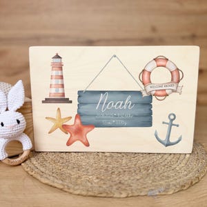A light wooden box with a nautical design on its lid, featuring a lighthouse, starfish, a lifebuoy with "WELCOME ABOARD" text, an anchor, and a hanging sign with "Noah" and birth details.