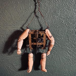 May include: A vintage, steampunk-style art piece featuring a clock mechanism body with attached wooden doll arms and legs. The arms and legs are painted in a flesh tone, and the clock mechanism is made of brass. The piece is suspended by a wire.