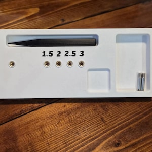 May include: A white rectangular tool with a black blade, labeled with the numbers 1.5, 2, 2.5, and 3. It has several small holes and a small compartment. Two silver screws are in a separate compartment. The tool is on a wooden surface.