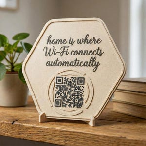 Könnte beinhalten: Ein sechseckiges Holzschild mit dem Text "home is where Wi-Fi connects automatically" und einem QR-Code. Das Schild steht auf einem Holztisch mit Büchern und einer Topfpflanze im Hintergrund.