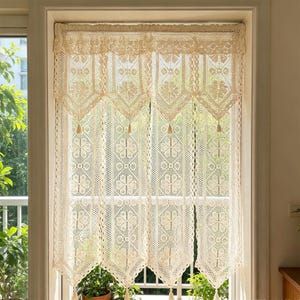 May include: Cream-colored, crocheted window curtain. The curtain features a floral and geometric pattern with a scalloped valance and tassels. The curtain hangs in front of a window with greenery visible outside.