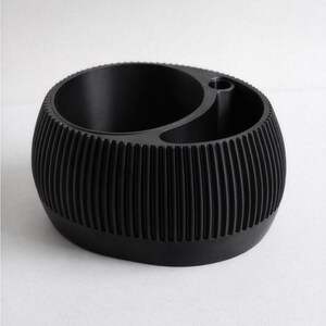 May include: A black desk tidy with a ribbed exterior. The organiser has two main compartments and a small central hole. The design is modern and minimalist, suitable for holding pens, pencils, or other stationery items.