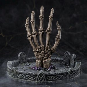 Può includere: Una scultura di mano scheletrica grigia e testurizzata che emerge da una base circolare in pietra. La base presenta marcatori scolpiti simili a lapidi e intricati motivi a intreccio lungo il bordo, con piccoli cristalli viola.