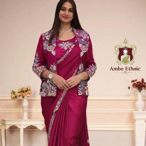 May include: A magenta saree and matching jacket with floral embroidery. The jacket has a round neckline and three-quarter sleeves. The saree has a decorative border and is draped over the body. The logo "Ambe Ethnic" is visible.